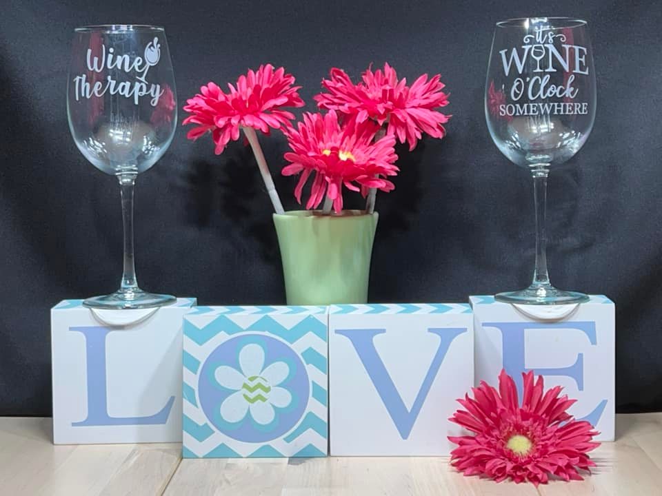 two wine glasses that say wine therapy and its wine o'clock somewhere. They are standing on top of blocks that spell out love with flowers in between them