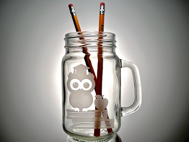 mason jar mug that have two books, an apple and owl with graduation cap on top and two pencils in the mug 