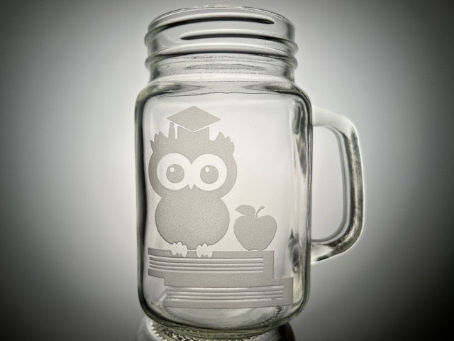 mason jar mug that have two books, an apple and owl with graduation cap on top 